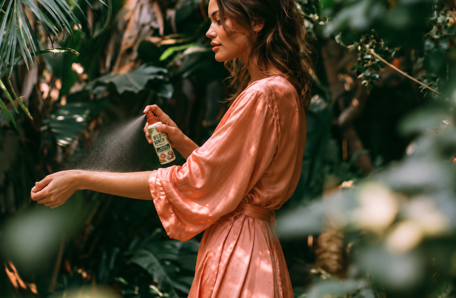 A woman in a coral dress spraying Wild Bunch Naturals repellent onto her bare forearm in a lush garden